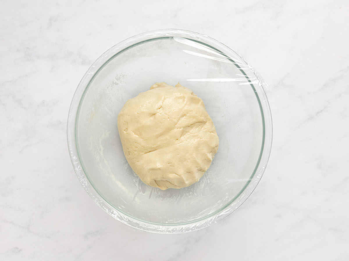 This is an overhead horizontal image of a glass bowl filled with a ball of dough. The bowl sits on a white marble counter. The bowl is covered in plastic wrap.