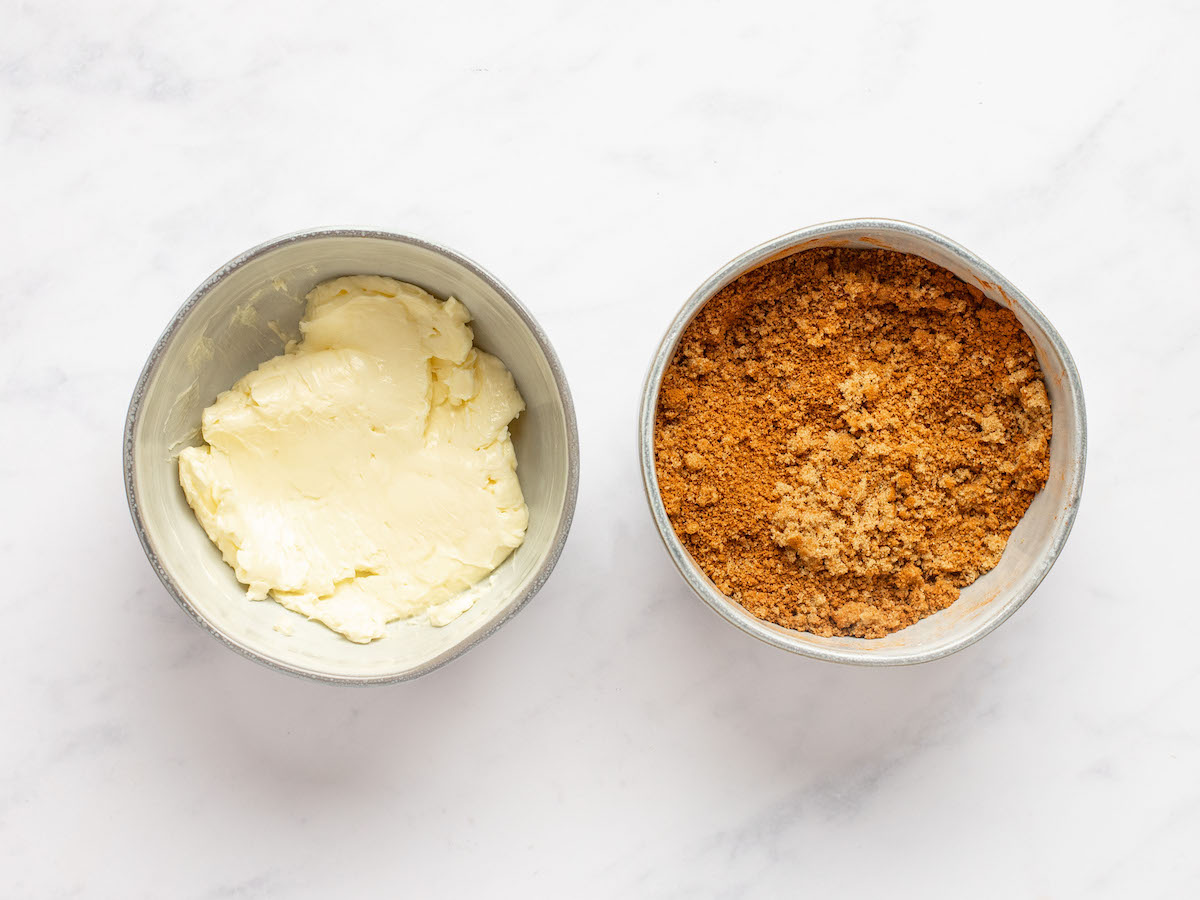 This is an overhead horizontal image of two small white bowls on a white marble surface. The bowl to the left has softened butter in it. The bowl to the right has cinnamon sugar in it. 