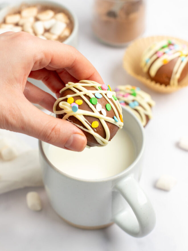 This is a vertical image of a hand coming in from the left side of the image holding a hot cocoa bomb over a white mug filled with milk. The hot chocolate bomb is drizzled with white chocolate and topped with rainbow sprinkles. The mug sits on a white marble surface with more mini marshmallows around it, a mug with hot chocolate and marshmallows and a hot chocolate bomb are blurred in the background.