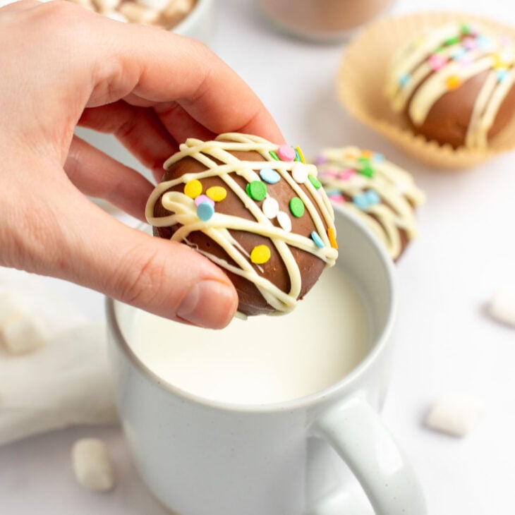 This is a vertical image of a hand coming in from the left side of the image holding a hot cocoa bomb over a white mug filled with milk. The hot chocolate bomb is drizzled with white chocolate and topped with rainbow sprinkles. The mug sits on a white marble surface with more mini marshmallows around it, a mug with hot chocolate and marshmallows and a hot chocolate bomb are blurred in the background.