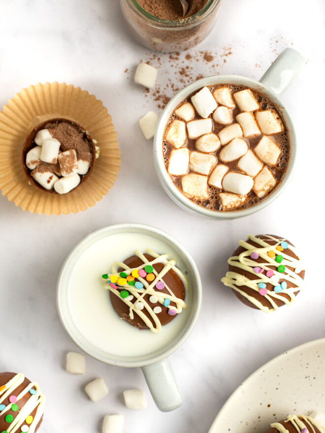 This is an overhead vertical image of a white marble surface with a white mug with hot chocolate and mini marshmallows, a mug with milk and a hot chocolate bomb floating on top and more hot chocolate bombs are on the marble counter around the two mugs.