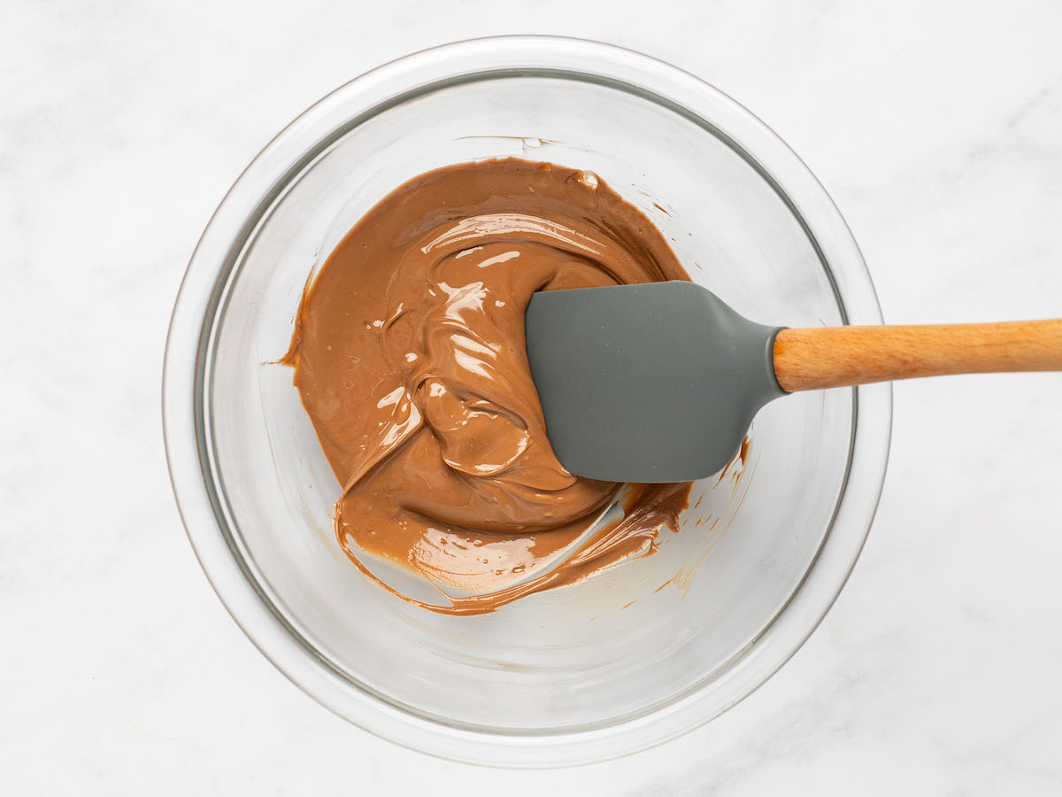 This is an overhead horizontal image of a glass bowl on a white marble surface. In the bowl is melted milk chocolate. A grey rubber spatula with a wooden handle is dipping into the chocolate and leaning against the right side of the bowl.