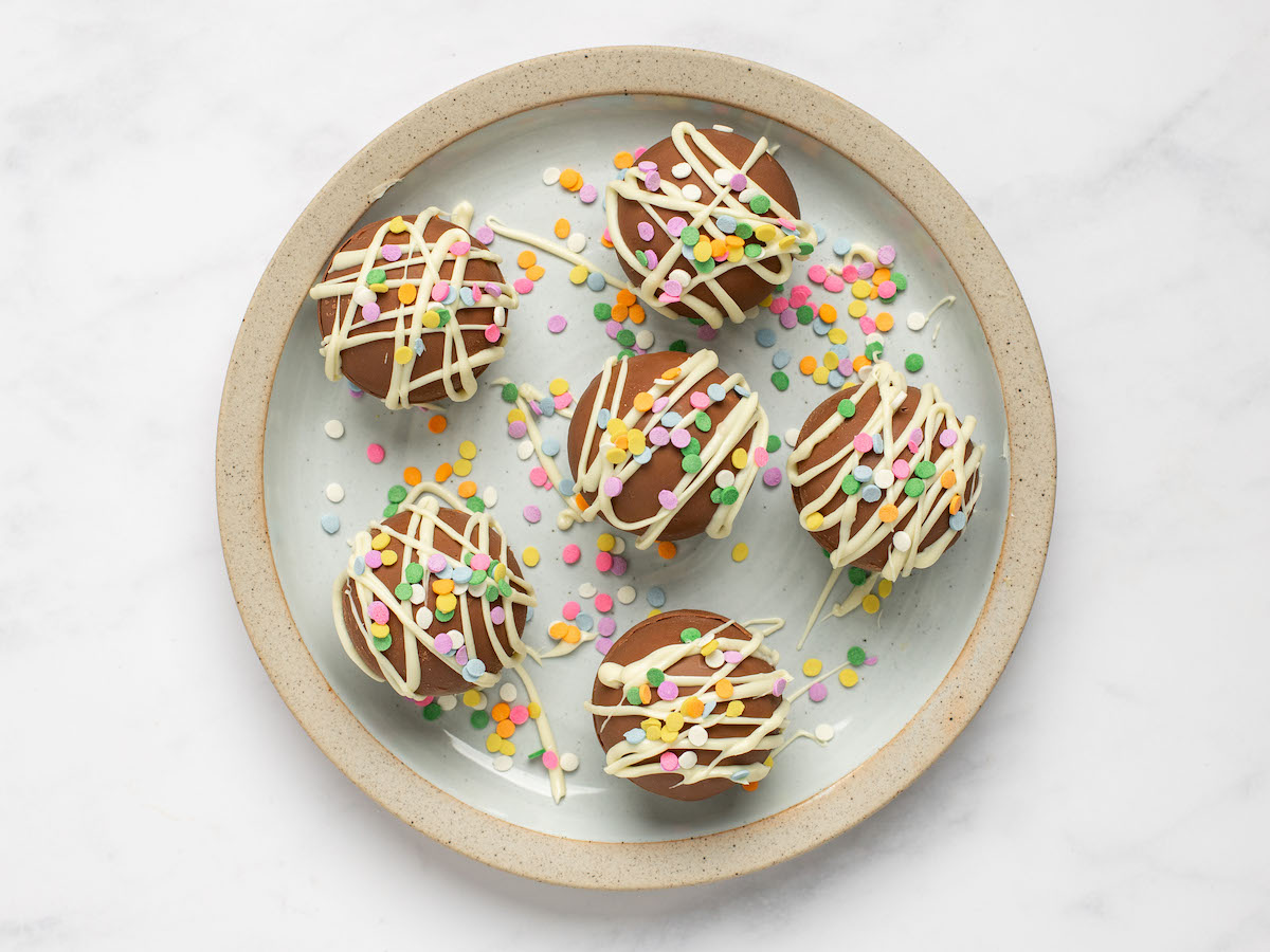 This is an overhead horizontal image of a white plate with a thick speckled beige rim sitting on a white marble surface. On the plate are six chocolate spheres drizzled with white chocolate and sprinkled with rainbow circle sprinkles. More sprinkles have fallen around the chocolate bombs.
