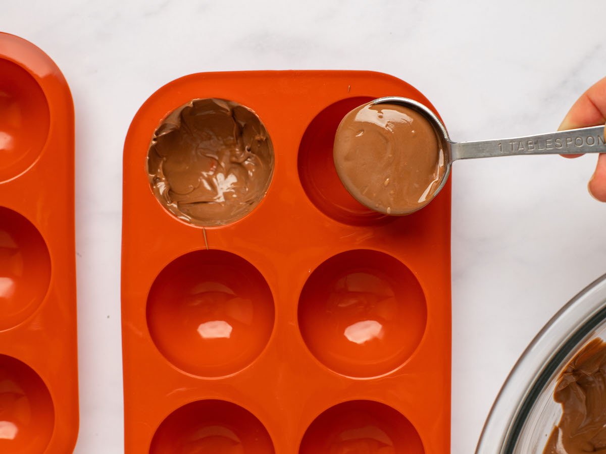This is an overhead horizontal image of a red silicone half-sphere mold on a white marble surface. A hand is coming in from the right side of the image holding a measuring spoon filled with melted chocolate. Melted chocolate has already been spread across the inside of one of the half-spheres. A glass bowl of melted chocolate is in the bottom right corner of the image.