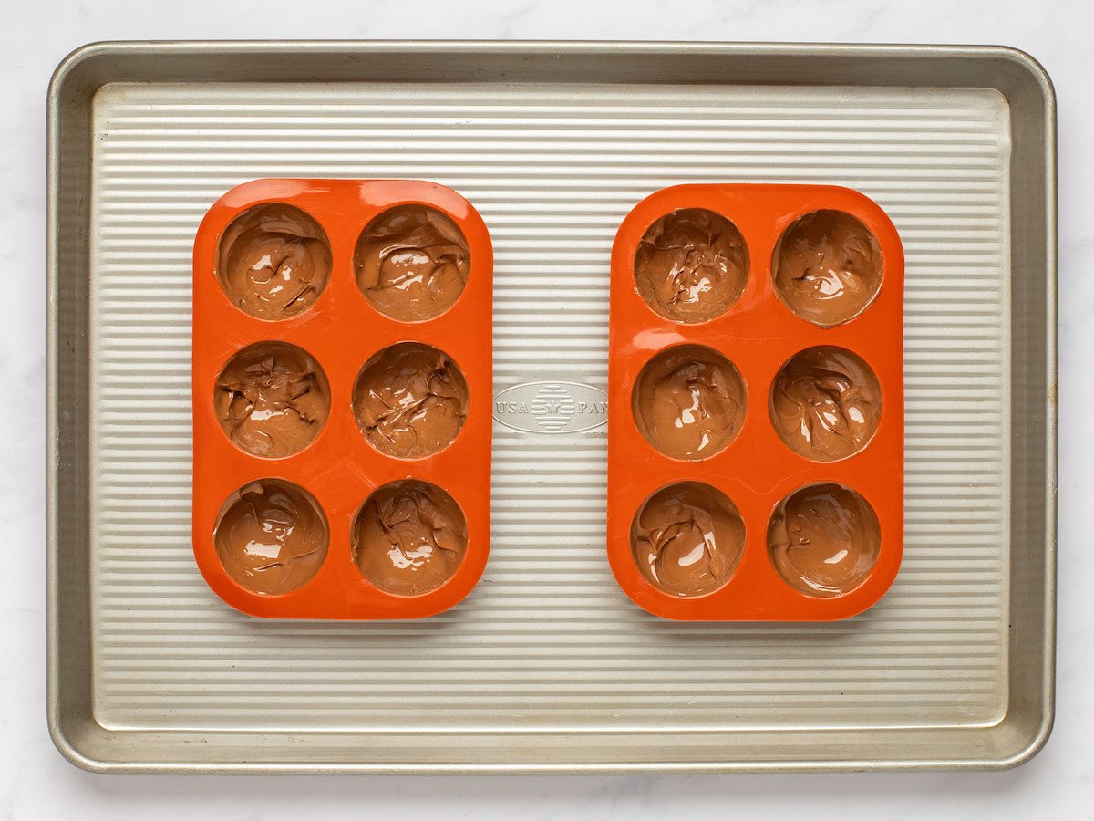This is an overhead horizontal image of a rimmed baking sheet on a white marble surface. On the pan are two red silicone hot chocolate bomb molds filled with melted chocolate.