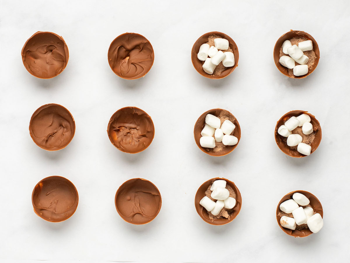 This is an overhead horizontal image of twelve half-spheres sitting on a white marble surface. On the left half of the image are six filled with hot chocolate mix and mini marshmallows. Six chocolate semi-spheres are to the left side of the image.