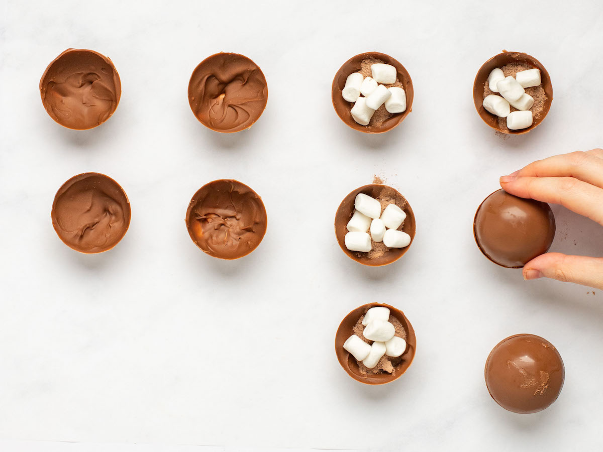 This is an overhead horizontal image of twelve half-spheres sitting on a white marble surface. On the left half of the image are six filled with hot chocolate mix and mini marshmallows. A hand is coming to the right side of the image placing the top half of the chocolate sphere on top. Four chocolate semi-spheres are to the left side of the image.