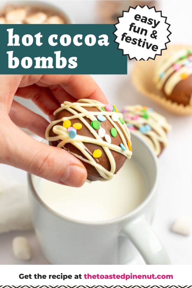 This is a vertical image of a hand coming in from the left side of the image holding a hot cocoa bomb over a white mug filled with milk. The hot chocolate bomb is drizzled with white chocolate and topped with rainbow sprinkles. The mug sits on a white marble surface with more mini marshmallows around it, a mug with hot chocolate and marshmallows and a hot chocolate bomb are blurred in the background. Text overlay reads "easy fun & festive hot cocoa bombs."