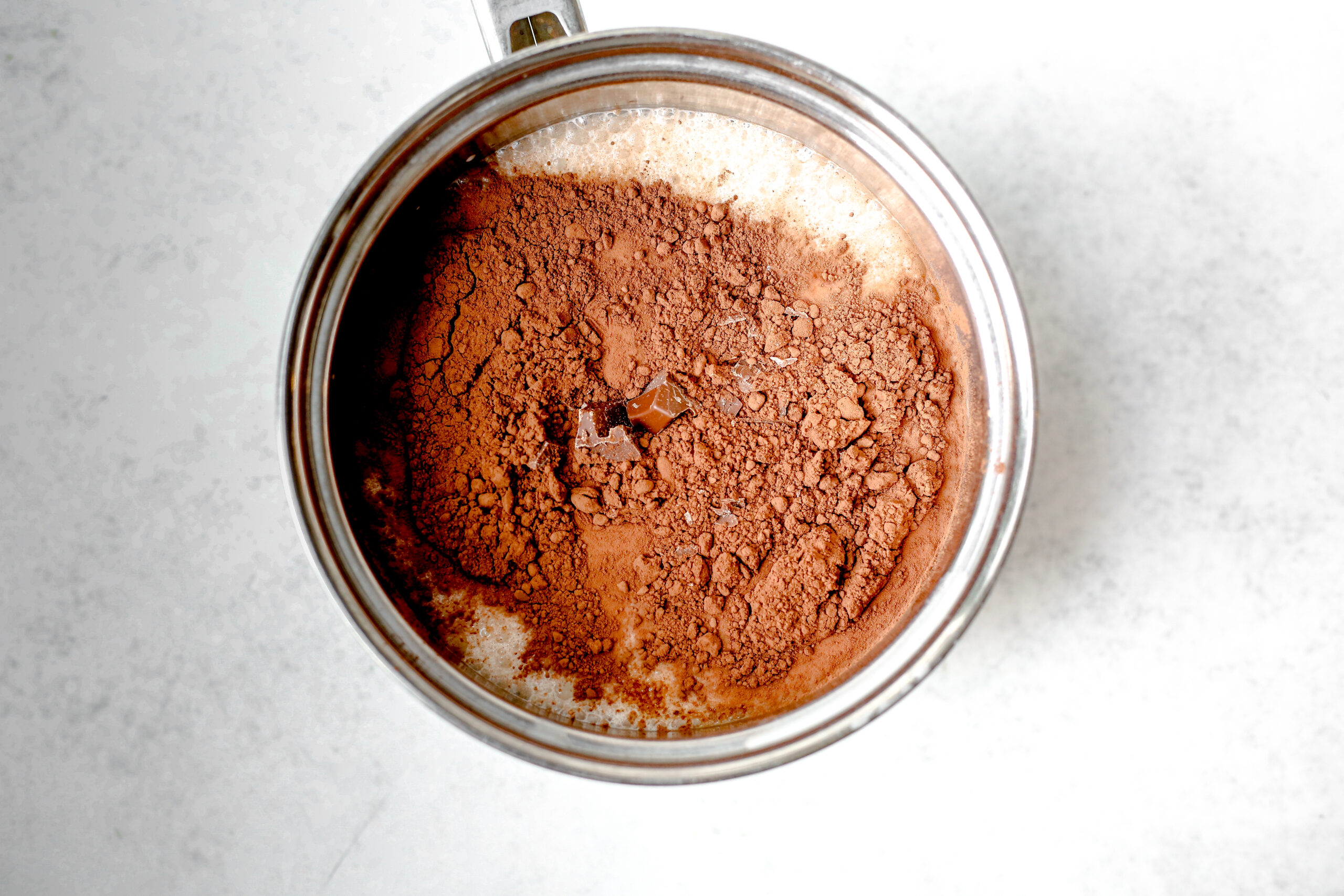 This is an overhead horizontal image of a silver saucepan with milk, cocoa powder and a few visible pieces of chopped chocolate in the center. The pot sits on a light grey surface.