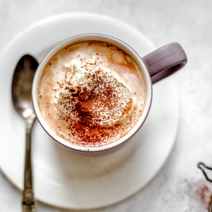This is an overhead horizontal image of a purple/grey mug on a small white plate. In the mug is hot chocolate with whipped cream and a sprinkle of cocoa powder. A silver spoon is on the plate next to the mug. The plate sits on a light grey surface.