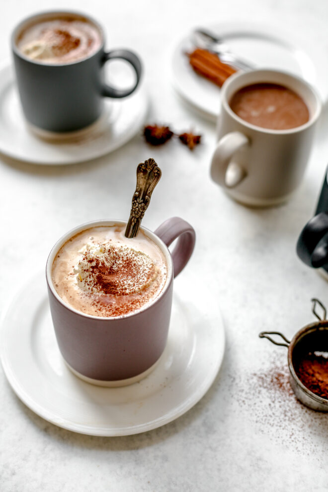 This is a vertical image looking at a purple mug filled with hot chocolate and whipped cream. The mug sits on a small white plate on a light grey surface. More mugs, one dark grey and one beige are blurred behind the purple mug in focus. Stars of anise and cocoa powder are around the mug on the grey surface.