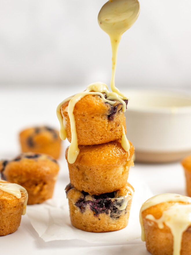 This is a vertical image looking at a stack of three mini blueberry muffins the side. The muffins sit on a small white piece of parchment paper on a white marble counter. A small white bowl and more mini blueberry muffins are blurred in the background behind the stack. A spoon with orange glaze is coming from the top center of the image and drizzling the glaze onto the muffin stack.