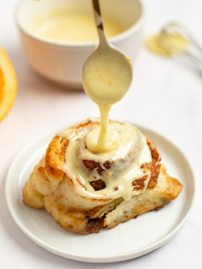 This is a vertical image looking at a cinnamon roll from the side. The cinnamon roll sits on a small white plate on a white marble counter. A small white bowl with orange glaze, a whisk, and a piece of an orange is blurred in the background behind the plate. A spoon with orange glaze is coming from the top center of the image and drizzling the glaze onto the cinnamon roll.