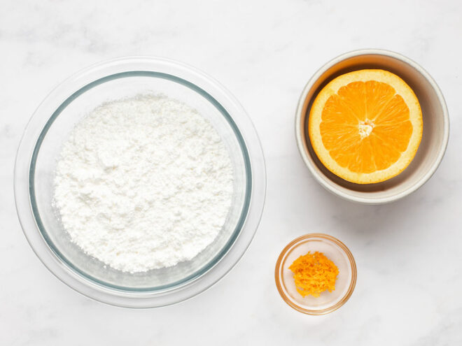 This is an overhead horizontal image of three bowls on a white marble surface. The large bowl to the left is glass and has powdered sugar in it. A smaller bowl with half an orange is to the top right of the image and the smallest bowl is to the bottom right of the image and is made of glass and has zest in it.