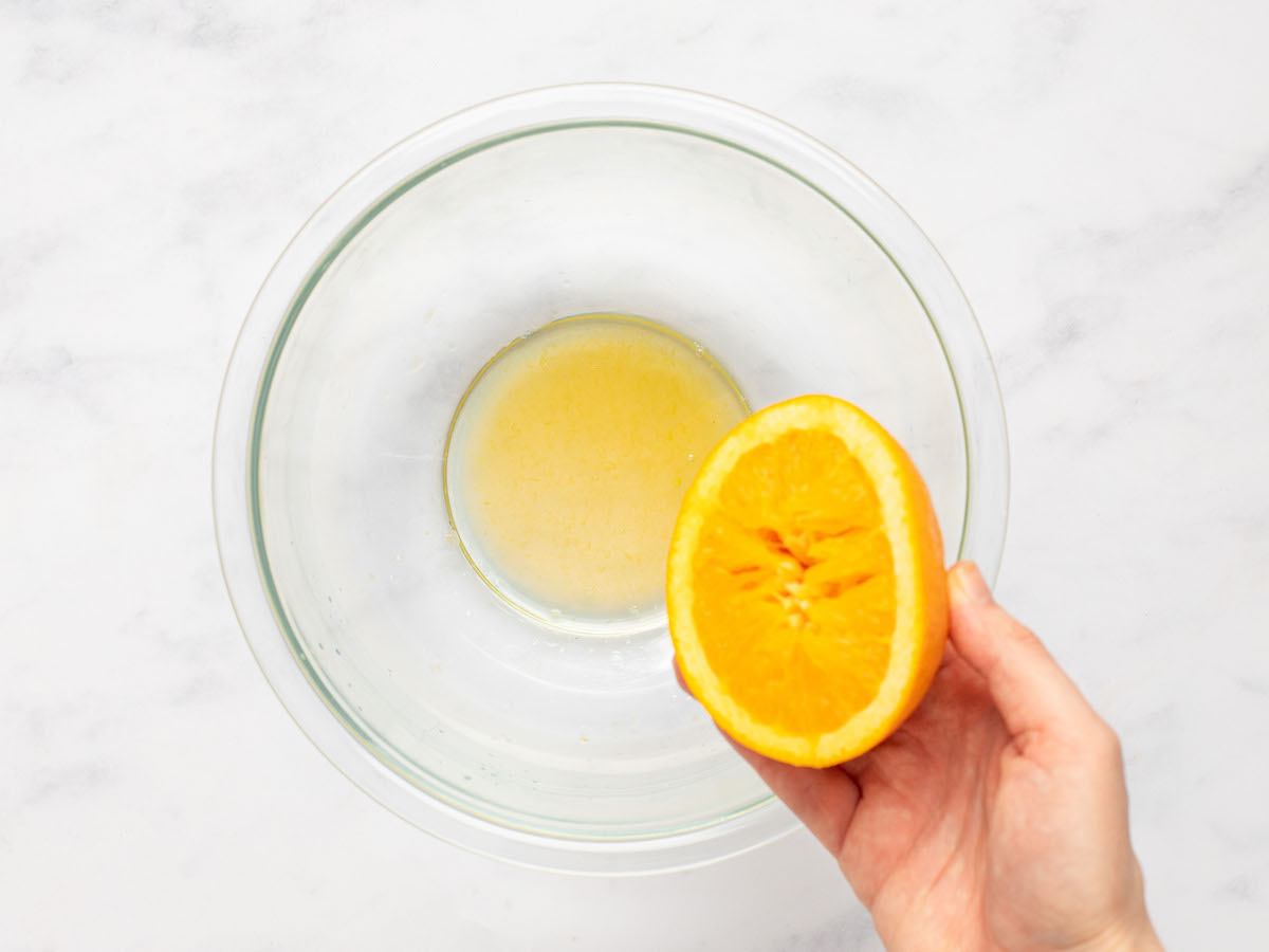 This is an overhead horizontal image of a glass bowl with orange juice in it. The glass bowl sits on a white marble counter. A hand is coming in from the bottom right corner of the image and is holding half of an orange.