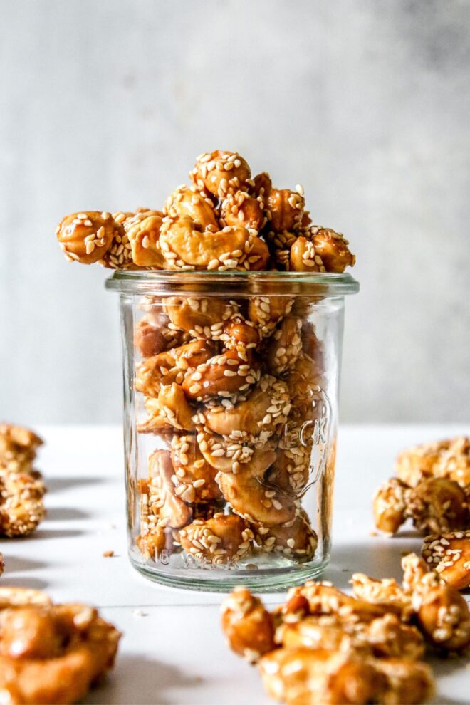 This is a vertical image looking at a glass jar from the side. In the jar are cashews with a deep honey color and coated with sesame seeds. The jar sits on a white surface with more clusters all around.