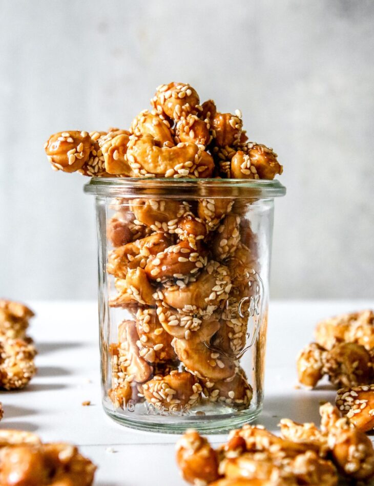 This is a vertical image looking at a glass jar from the side. In the jar are cashews with a deep honey color and coated with sesame seeds. The jar sits on a white surface with more clusters all around.