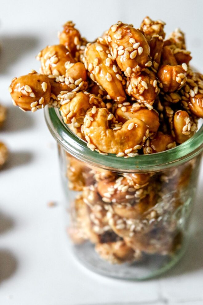 This is a vertical image looking at a glass jar from an upper angle. In the jar are cashews with a deep, shiny honey color and coated with sesame seeds. The jar sits on a white surface with more clusters all around.