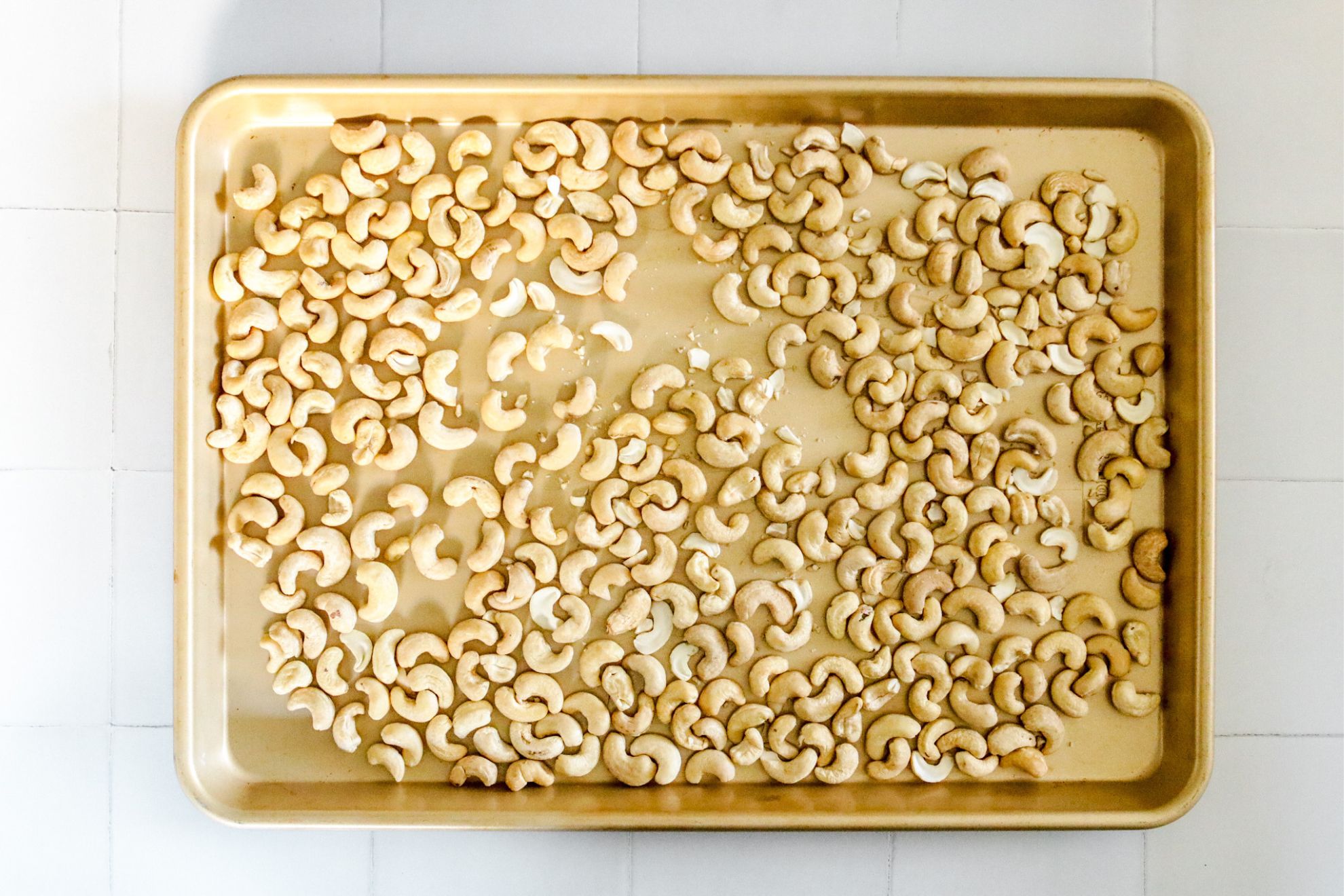 This is an overhead horizontal image of a gold rimmed baking sheet with raw cashews spread across it. The baking sheet sits on a white square tile surface.