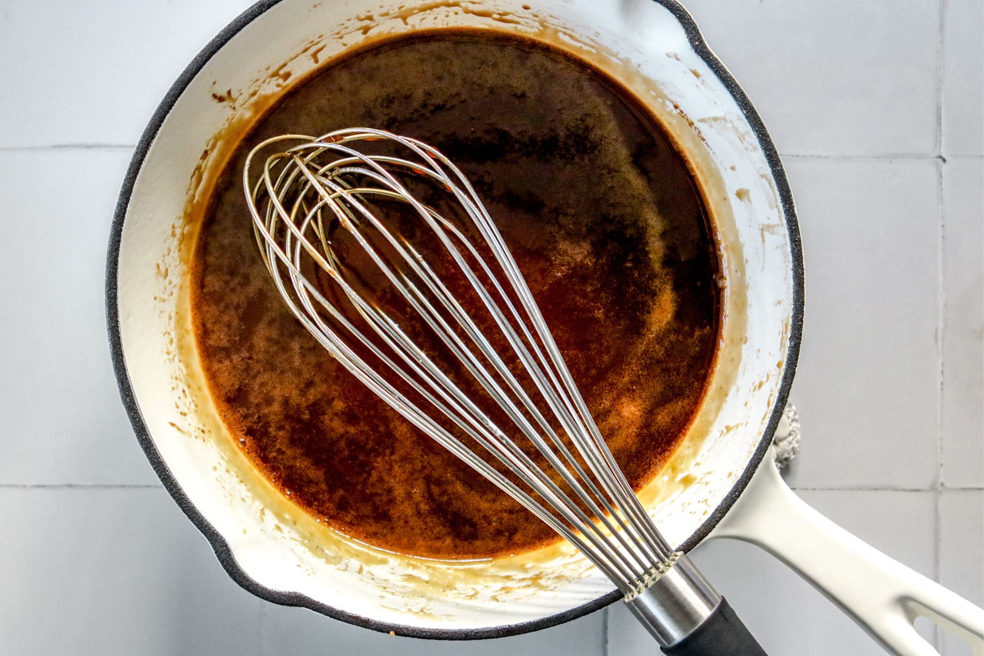 This is an overhead horizontal image of a small white saucepan with a caramel mixture. A silver whisk is in the caramel with the handle leaning against the rim and pointing to the bottom of the image. The pan sits on a white square tile surface.