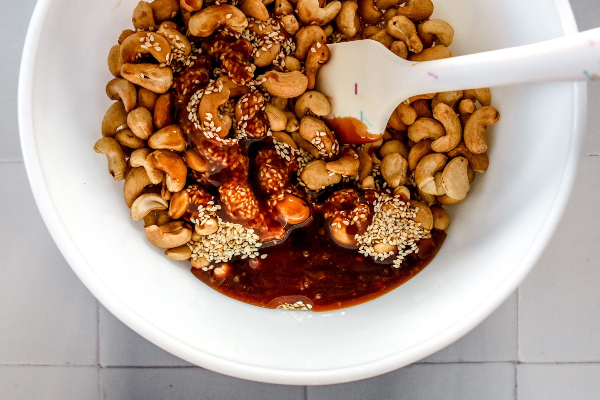 This is an overhead horizontal image of a large white bowl on a white tile surface. In the bowl are cashews, sesame seeds and a caramel sauce. A white rubber spatula is in the bowl with the handle leaning against the side of the bowl pointing to the top right of the image.