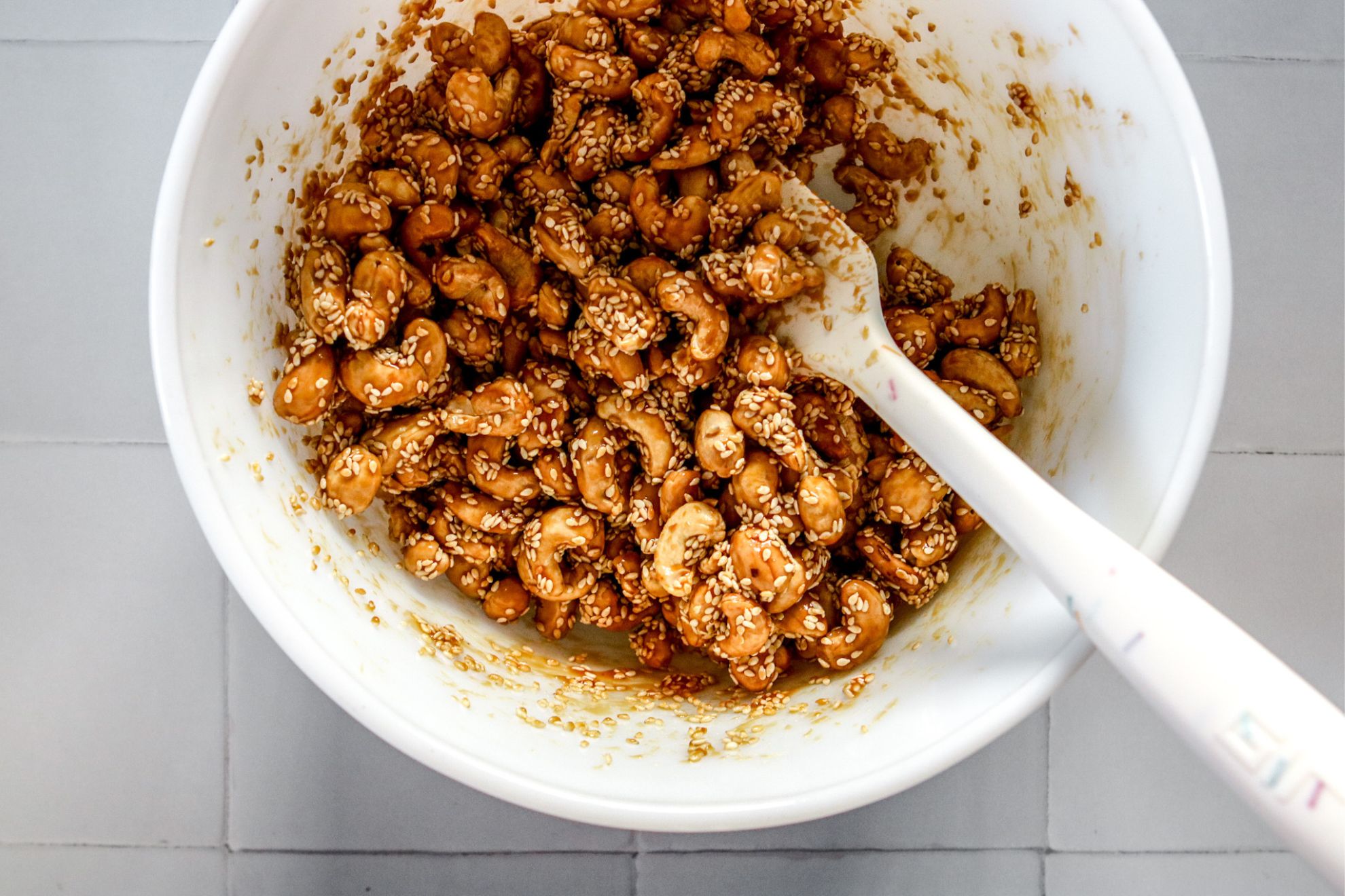 This is an overhead horizontal image of a large white bowl on a white square tile surface. In the bowl are cashews coated with sesame seeds and a caramel sauce. A white rubber spatula is in the bowl with the handle leaning against the side of the bowl pointing to the bottom right of the image.