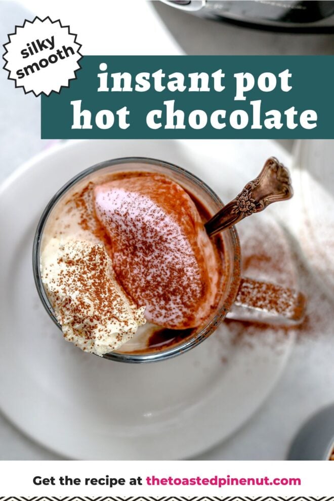 This is an overhead vertical image of a glass mug with hot cocoa and marshmallows on top. The mug is sprinkled with cocoa powder and sits on a small white plate. A vintage silver spoon dips into the mug and is sticking out of the mug. An instant pot is coming in from the top right corner of the image. Text overlay reads "silky smooth instant pot hot chocolate."