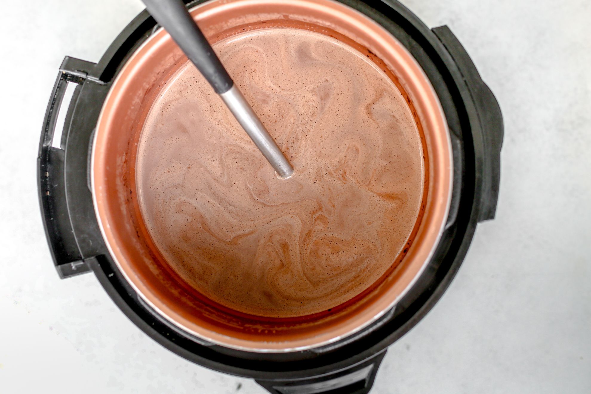 This is an overhead horizontal image looking into an instant pot with hot cocoa in it. A ladle is in the instant pot with the handle leaning against the left top side of it. The instant pot is on a light grey surface.
