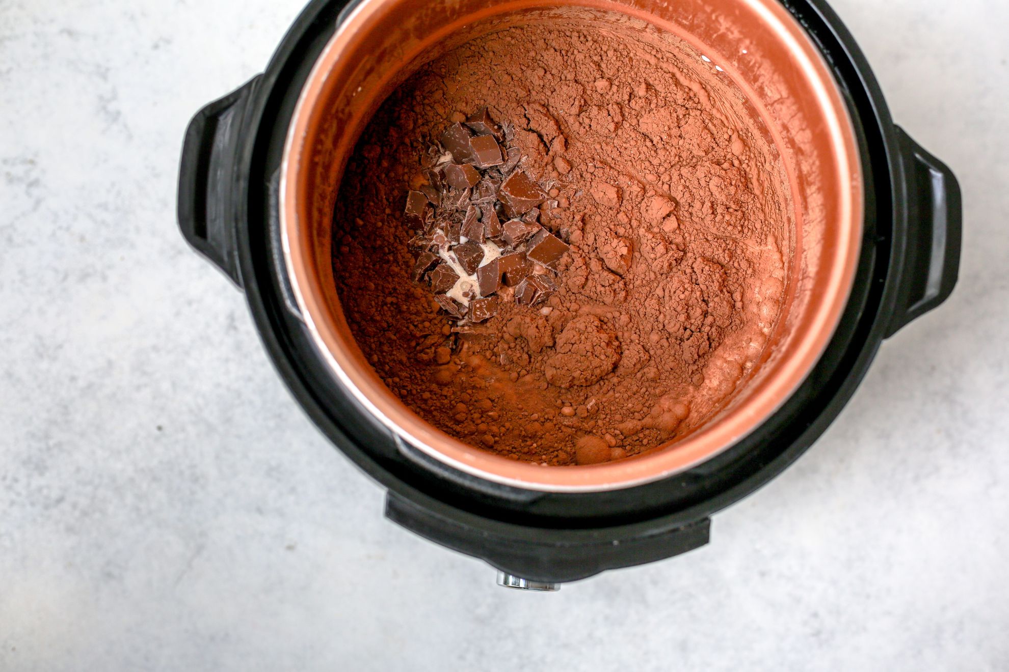This is an overhead horizontal image looking into an instant pot with cocoa powder, chopped chocolate and milk in it, not yet stirred or combined. The instant pot is on a light grey surface.