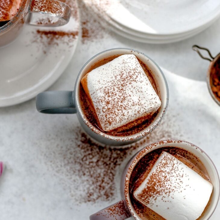 This is an overhead vertical image of two grey mugs on a light grey surface. in the mugs are hot cocoa with a large square marshmallow and a sprinkle of cocoa powder. To the top of the mugs is a stack of small white plates with silver spoons on it. Another mug with hot cocoa on a white plate is to the left top of the image.