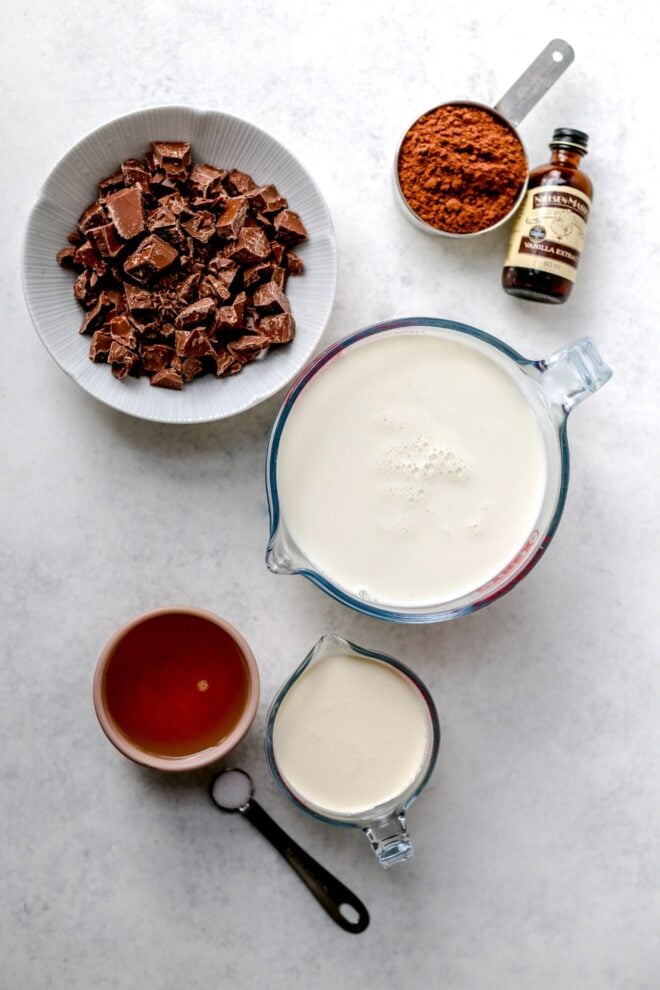 This is an overhead vertical image of ingredients on a light grey surface. There is a small white bowl with chopped chocolate, a measuring cup with cocoa powder, a small bottle of vanilla extract, a liquid measuring cup with milk and another smaller liquid measuring cup with cream. A small teaspoon of salt and a small bowl with an amber sweetener is in it.