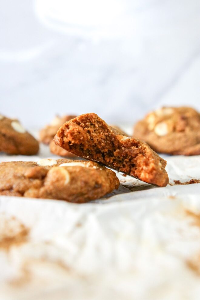 This is a vertical image looking at a cookie with a bite taken out of it from the side. The cookie leans against another cookie with white chocolate chips and more cookies are blurred in the background. All cookies sit on a white piece of parchment paper.