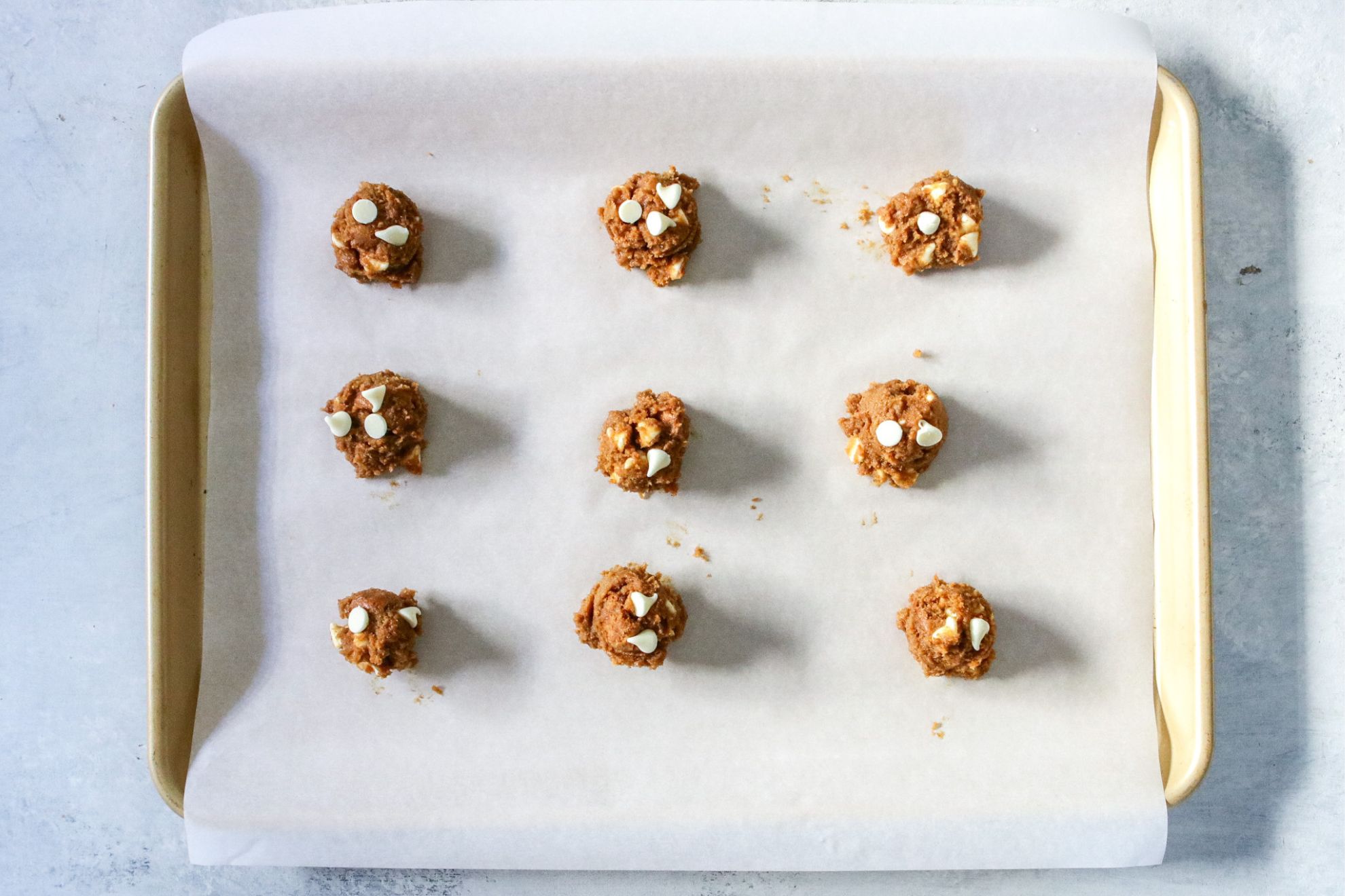 This is an overhead horizontal image of a gold rimmed baking sheet lined with white parchment paper. On the baking sheet is nine cookie dough balls topped with white chocolate chips. The baking sheet sits on a light grey surface.