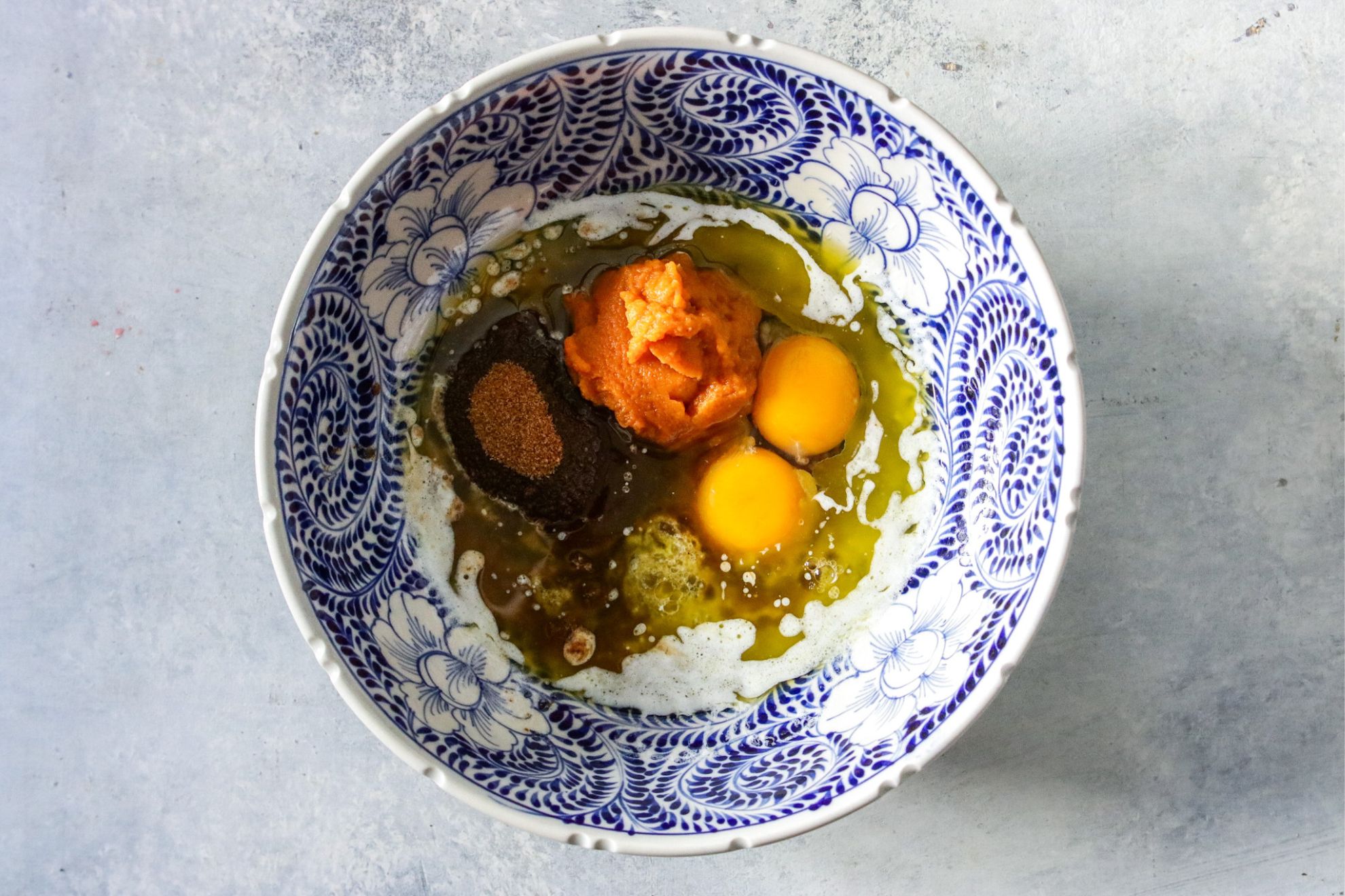 This is an overhead vertical image of a white and blue patterned bowl on a light grey surface. In the bowl are butter, eggs, pumpkin puree and coconut sugar.