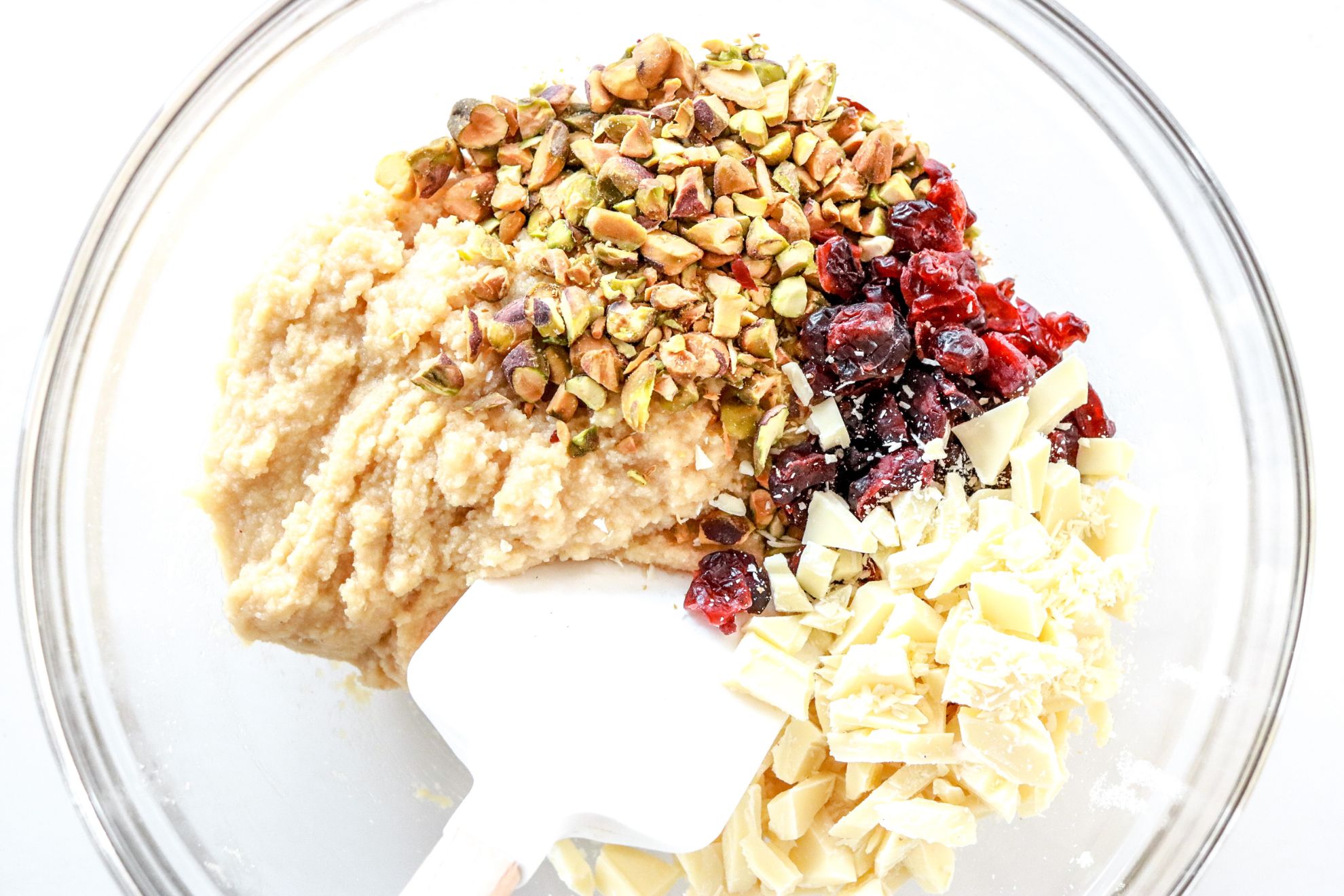 This is an overhead horizontal image of a glass bowl with raw cookie dough in it. On top of the dough are pistachio pieces, dried cranberries and white chocolate chunks. A spatula is dipping into the bowl. The bowl sits on a white surface.