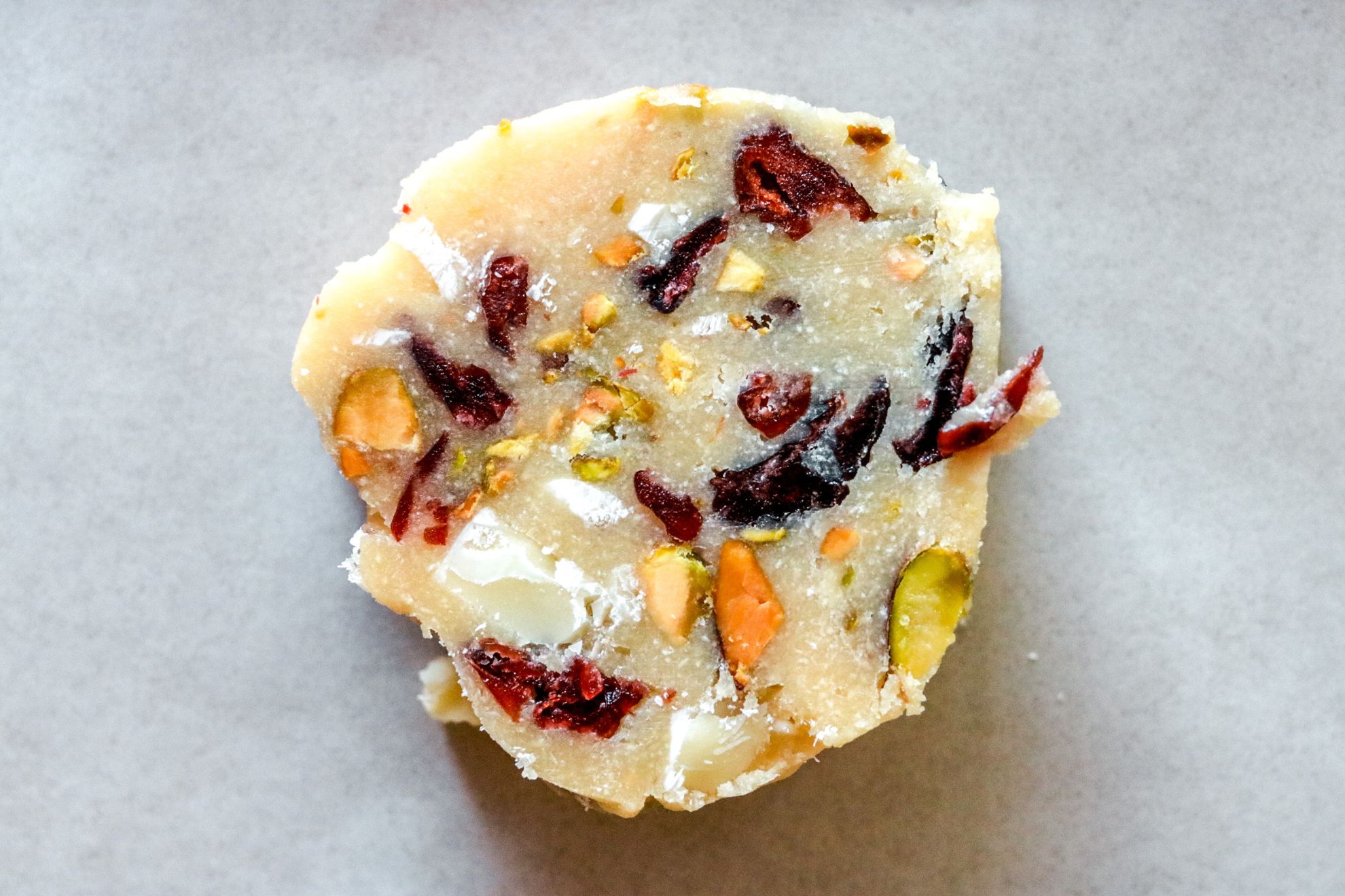 This is an overhead horizontal image of a close up of a sliced raw cookie dough circle. The cookie sits on a white piece of parchment paper and there are bits of dried cranberries, pistachios and white chocolate in it.