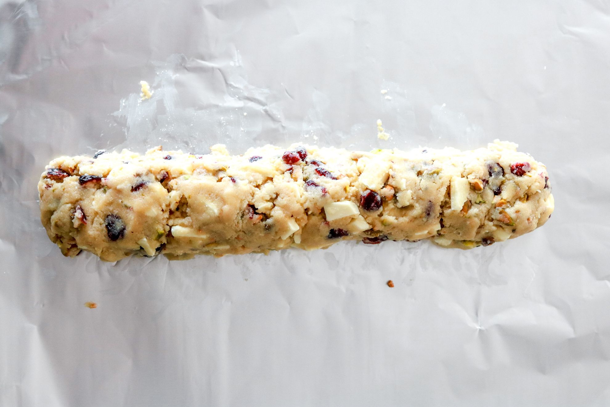This is an overhead horizontal image of a shortbread cookie dough log with dried cranberries, white chocolate and pistachio pieces in it. The log sits on a piece of tin foil.