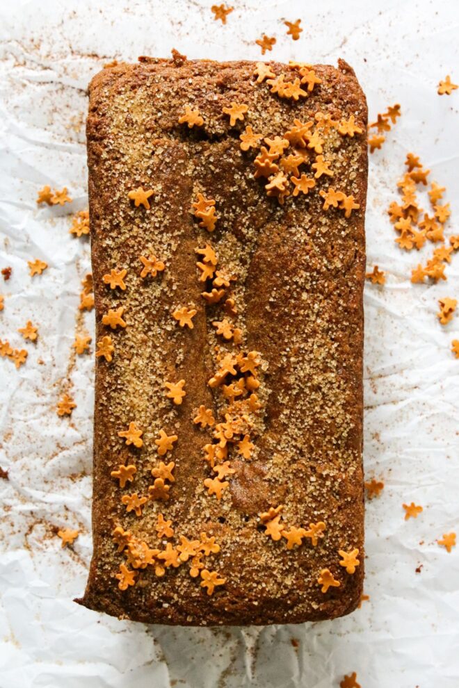 This is an overhead vertical image of a gingerbread loaf on a white piece of parchment paper. The loaf is topped with turbinado sugar and mini gingerbread men sprinkles.