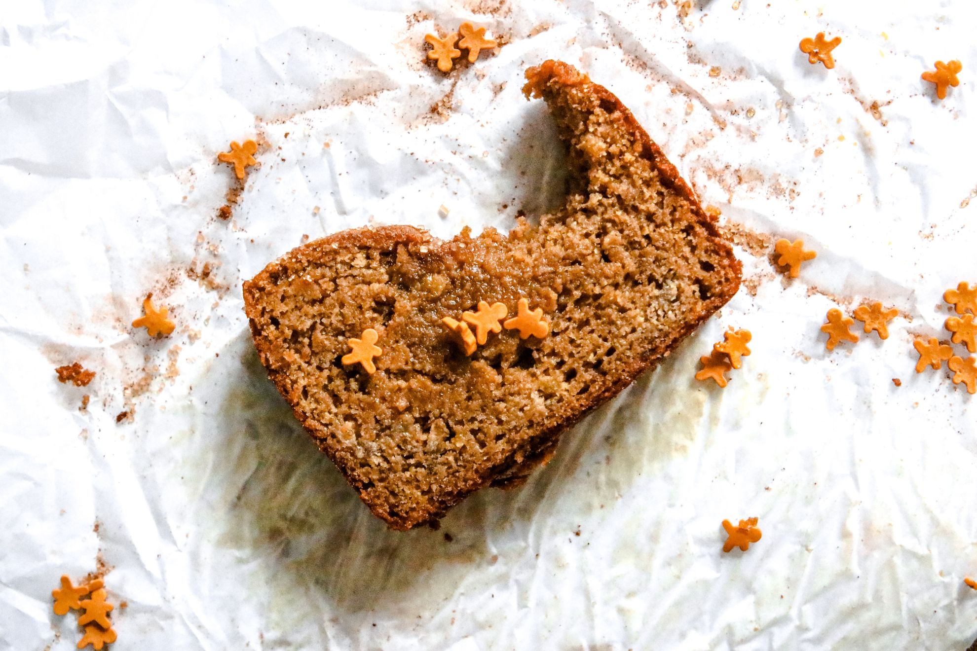 This is an overhead horizontal image of a slice of gingerbread banana bread with a bite taken out of it. The slice sits on a white piece of parchment paper and is sprinkled with mini gingerbread men sprinkles.