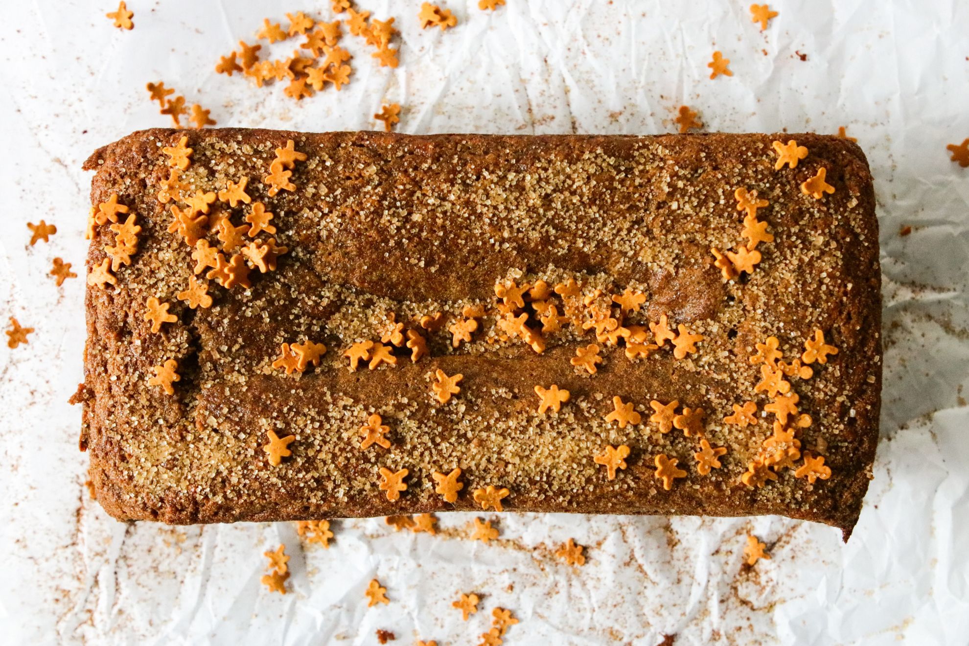 This is an overhead horizontal image of a gingerbread loaf on a white piece of parchment paper. The loaf is topped with turbinado sugar and mini gingerbread men sprinkles.