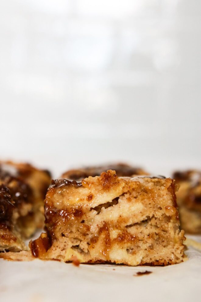This is a vertical side view of a piece of apple cake. The image hones in on the texture of the cake, revealing a moist texture with gooey cinnamon sugar on top and to the side. Noticeable in the cake are layers of thinly sliced apple. The piece of cake is on a white surface with a white background and more pieces of cake are blurred to the left and behind this piece in focus.