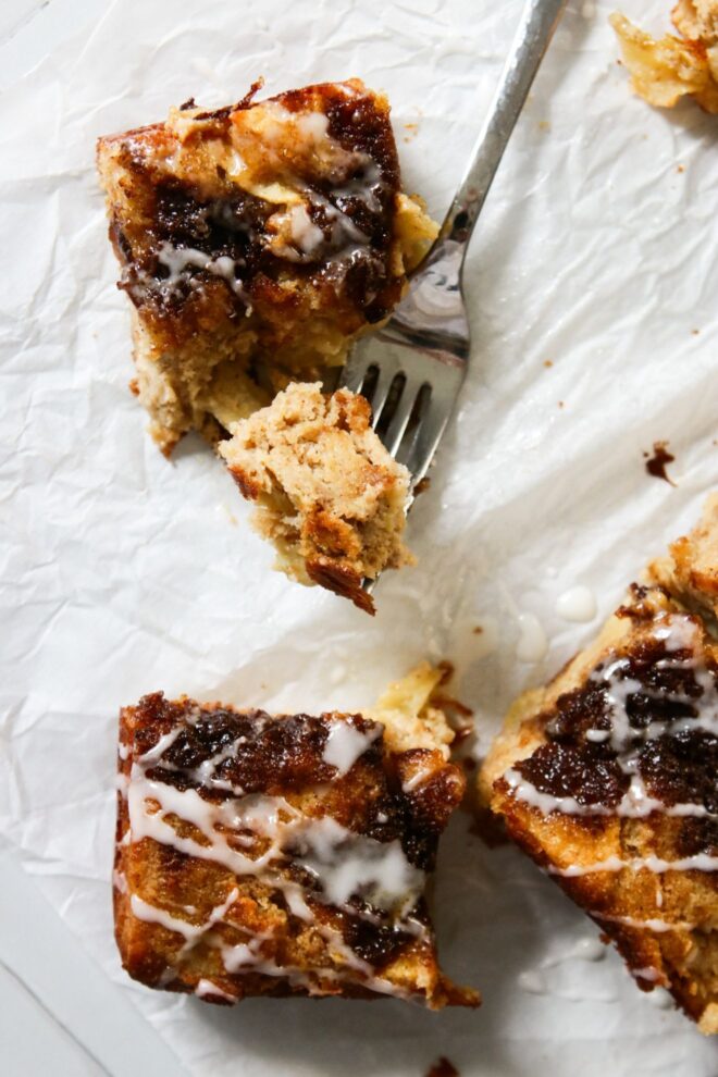 This is an overhead vertical image of three pieces of apple cake. The first slice is at the top left of the image and a fork is laying next to it with a bite on its prongs. the second and third square slices are to the bottom middle and bottom right of the image. All three square slices of cake have gooey cinnamon sugar on top and are drizzled with white icing. The cake pieces sit on a white piece of parchment paper.