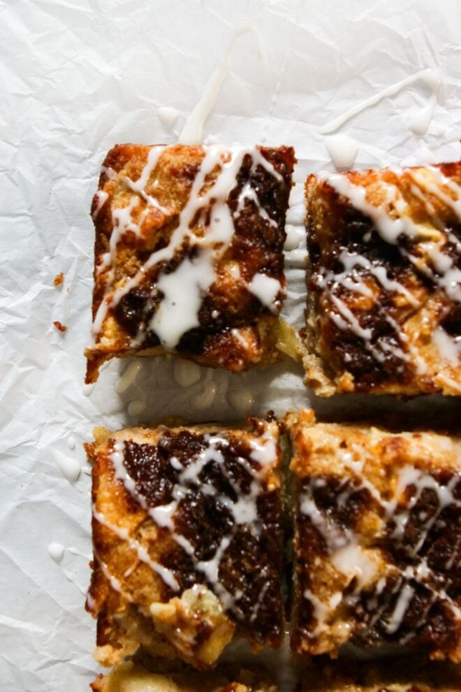 This is a vertical overhead image looking down onto apple cake cut in squares and drizzled with a white icing. The cake has puddles of cinnamon sugar on top. The cake sits on a crinkled white piece of parchment paper.