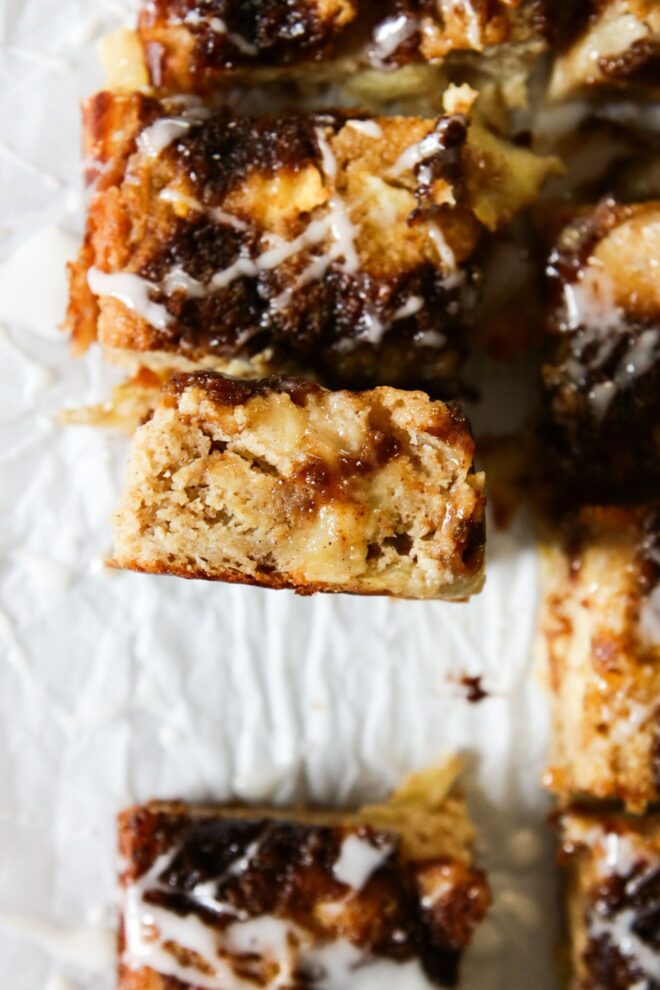 This is an overhead vertical image of a square of apple cake standing on its side to review the moist center. The cake has gooey cinnamon sugar on top. The cake slice is on a white piece of parchment paper with more cake squares to the top, right and bottom of this slice in focus.