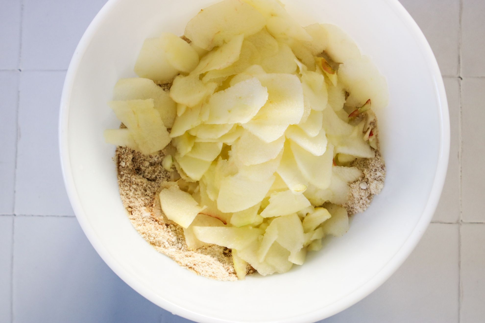 This is an overhead horizontal image of a large white bowl on a white square tile counter. In the bowl are a mix of dry ingredients with sliced apples on top.