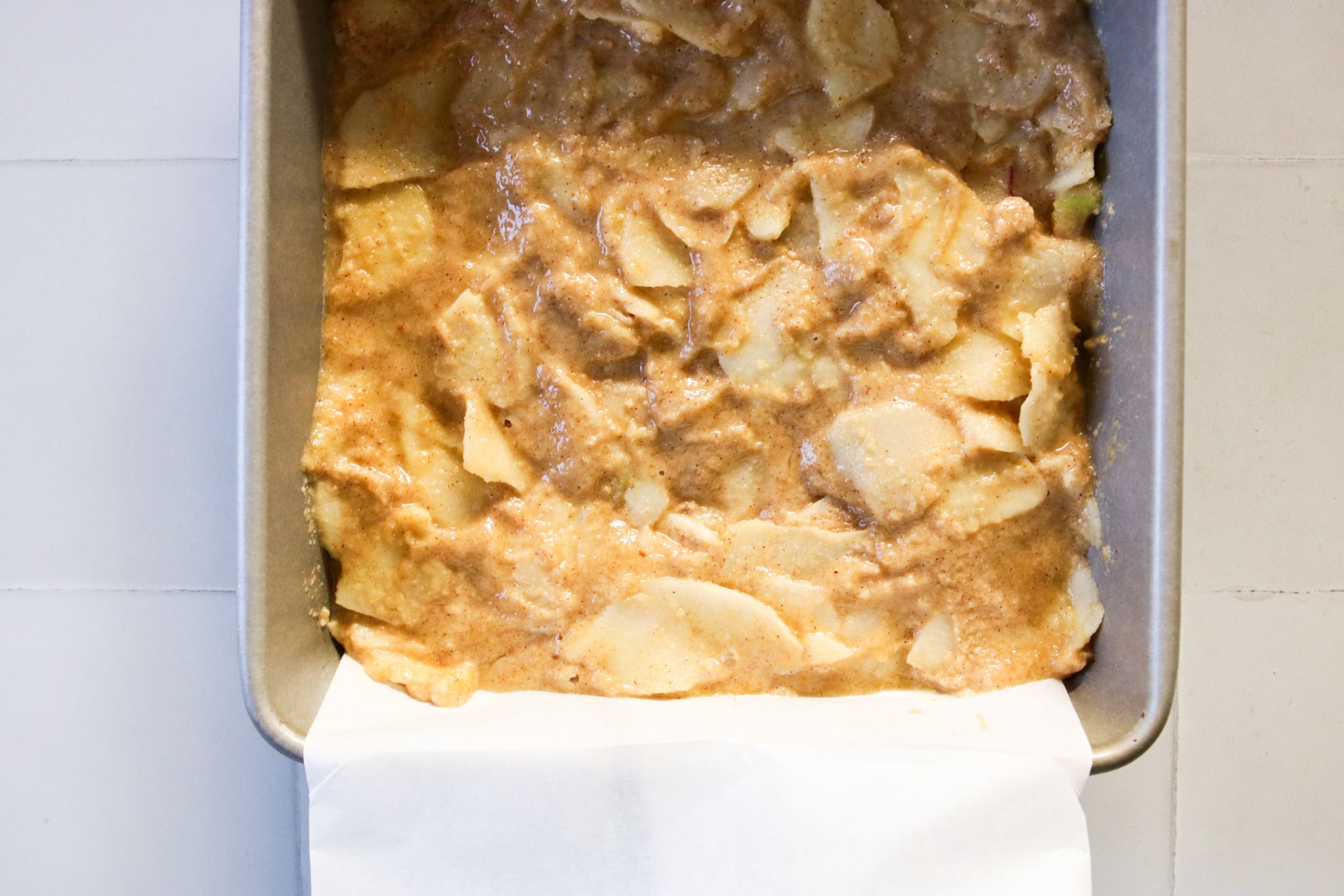 This is an overhead horizontal image of a silver square pan lined with parchment paper. In the pan is a raw cake batter with a lot of apple slices in it. The pan sits on a white square tile counter.