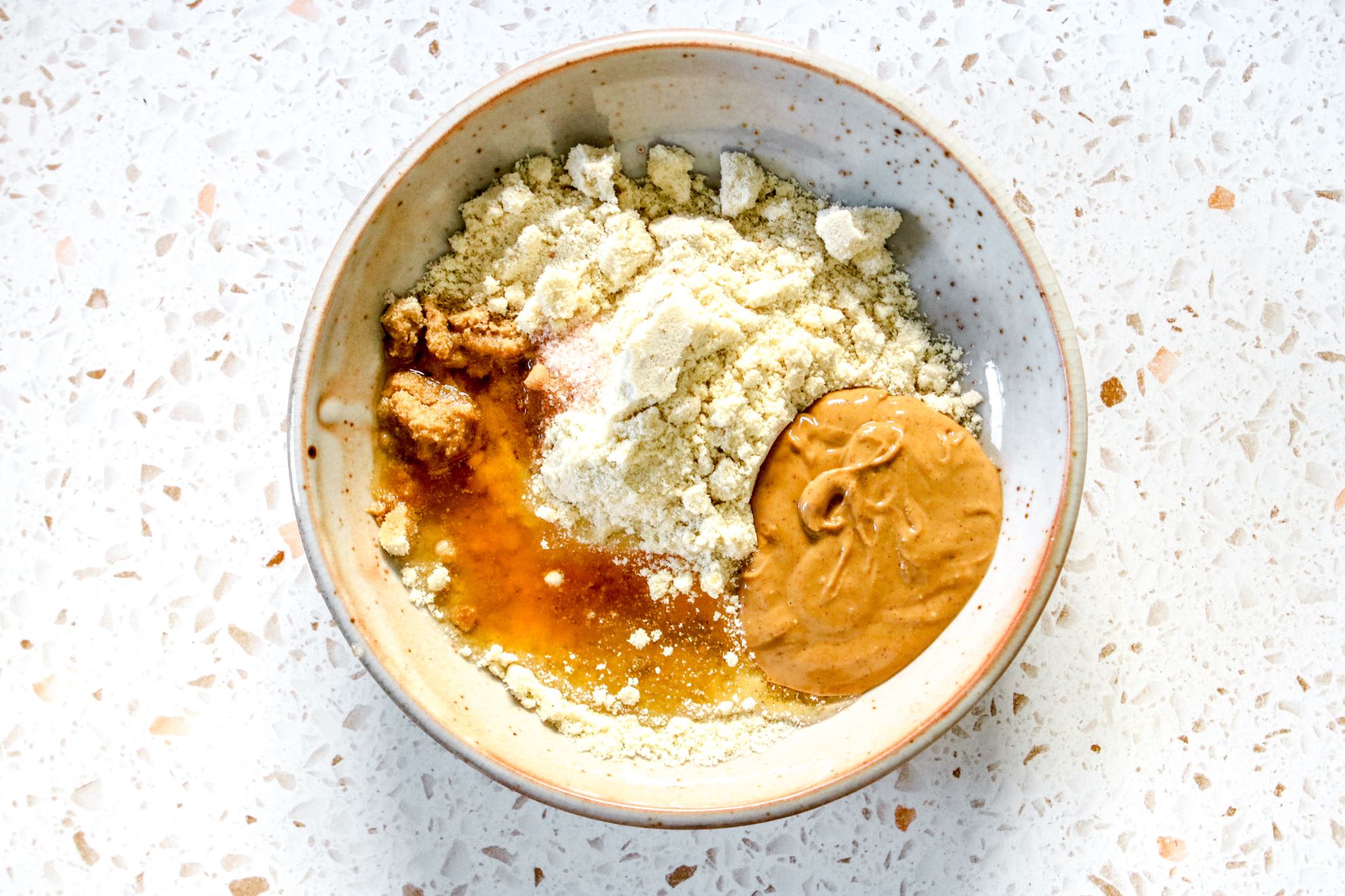 This is an overhead horizontal image of a bowl on a terrazzo surface. In the bowl is almond flour, peanut butter, agave nectar, salt, and vanilla extract.