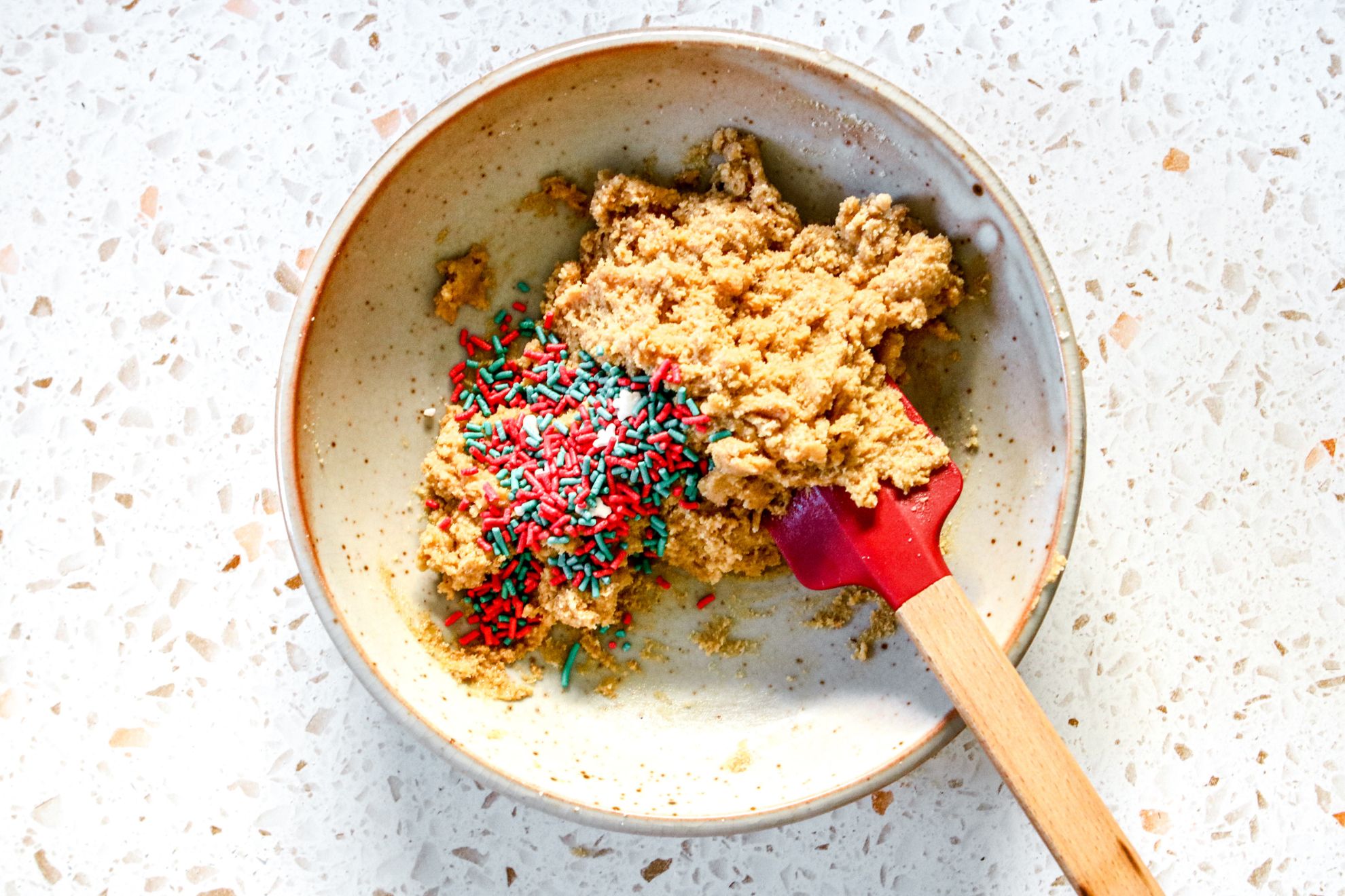 This is an overhead horizontal image of a bowl on a terrazzo surface. In the bowl is a raw cookie dough with red and green sprinkles piled to the left. A red spatula with a wooden handle is in the bowl leaning against the side and pointing to the bottom right corner of the image.