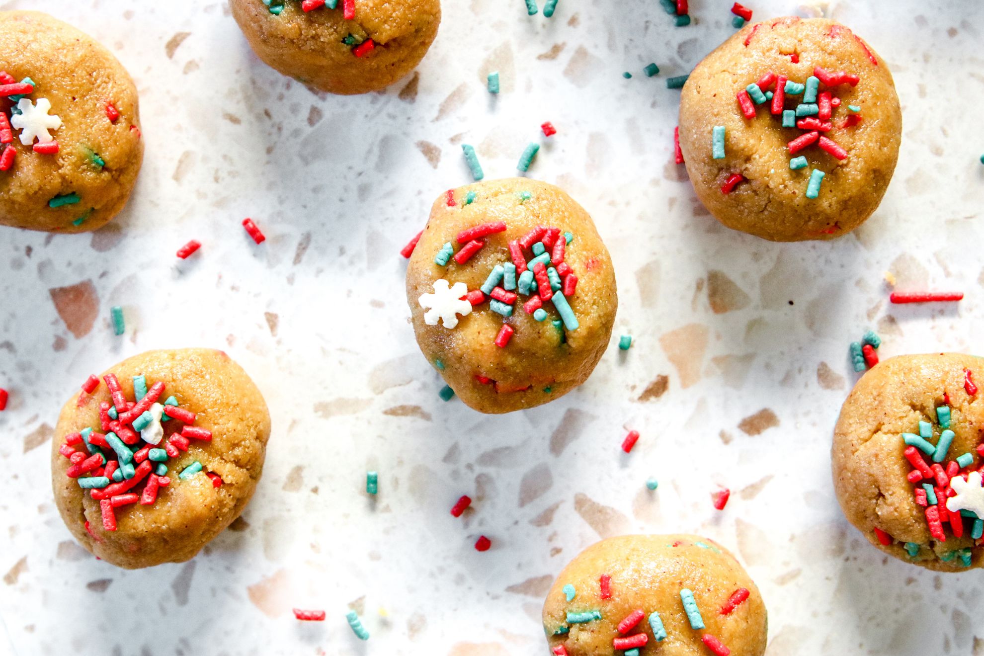 This is an overhead horizontal image of cookie dough balls on a white terrazzo surface. The balls have red green and snowflake sprinkles on top.