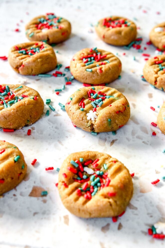 This is vertical image of cookies on a white terrazzo surface. The cookies have red green and snowflake sprinkles on top and the tops have been pressed down with the back of a fork. More sprinkles are on the surface around the cookies.